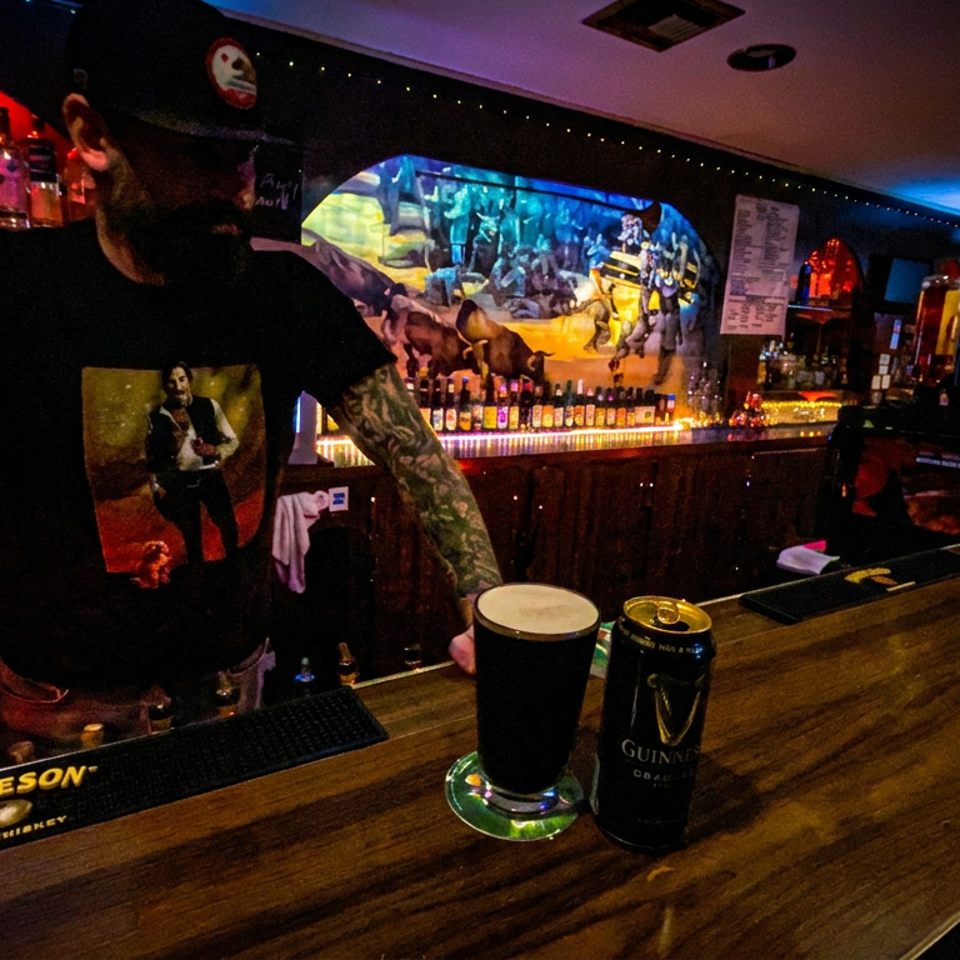 Tattooed bartender serves drinks behind a dimly lit bar stocked with liquor, featuring colorful artwork and Guinness and Jameson branding.