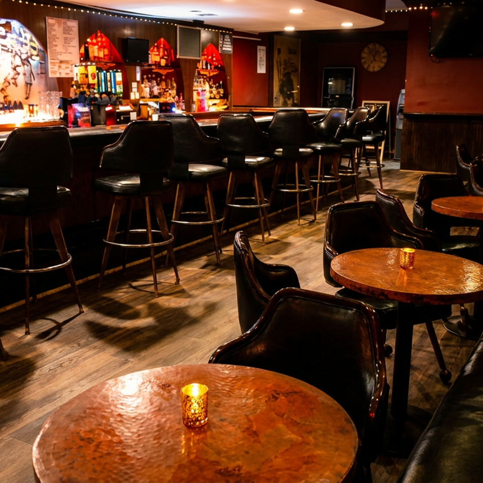 Dimly lit bar interior with leather stools, copper tables, candlelit seating, colorful liquor shelves, and mural artwork.